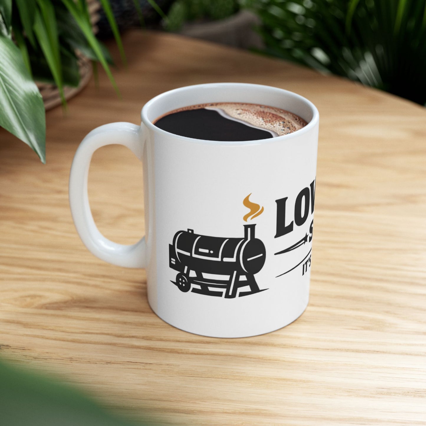 Low & Slow Society funny BBQ pitmaster mug, white ceramic, lifestyle shot on a wooden table beside houseplants, filled with black coffee, handle on the left