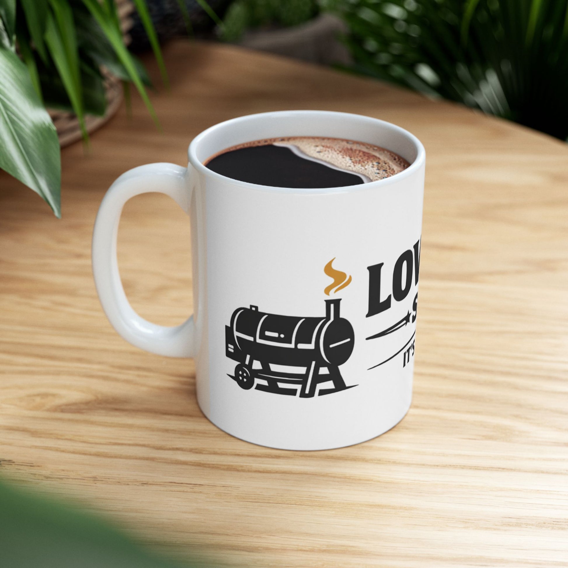 Low & Slow Society funny BBQ pitmaster mug, white ceramic, lifestyle shot on a wooden table beside houseplants, filled with black coffee, handle on the left