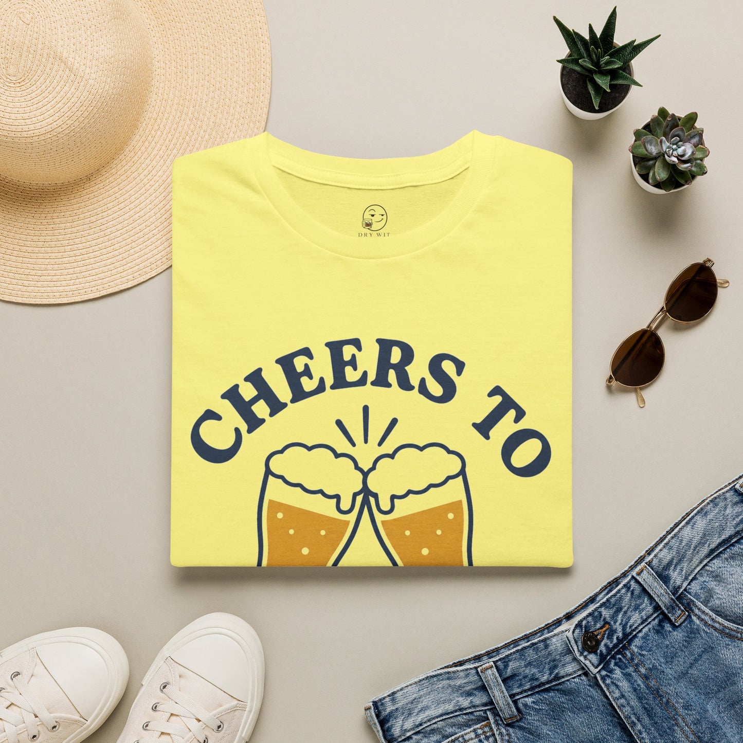Cheers to Lowering Expectations Shirt, Cornsilk pale yellow, folded flat lay styled with a straw hat, sunglasses, sneakers and denim