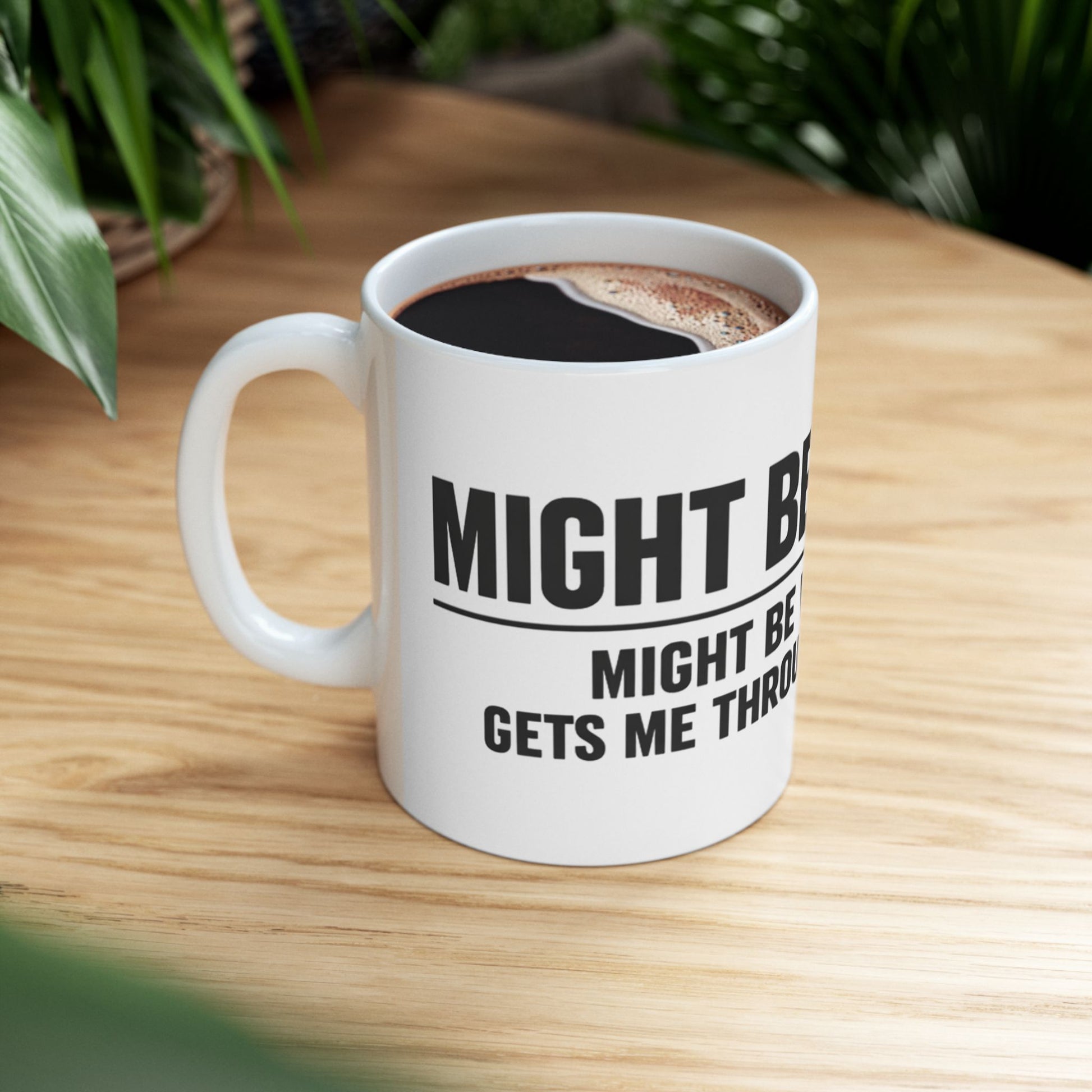Might Be Coffee Mug, white ceramic, lifestyle shot on a wooden table beside houseplants, filled with black coffee, handle on the left