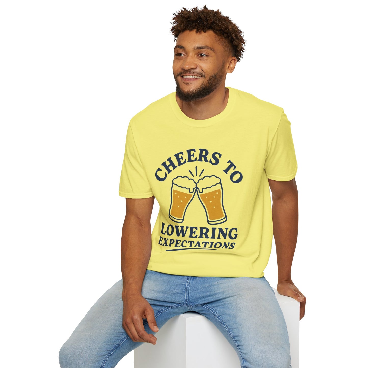 Cheers to Lowering Expectations Shirt, Cornsilk pale yellow, worn by a model seated on a white block