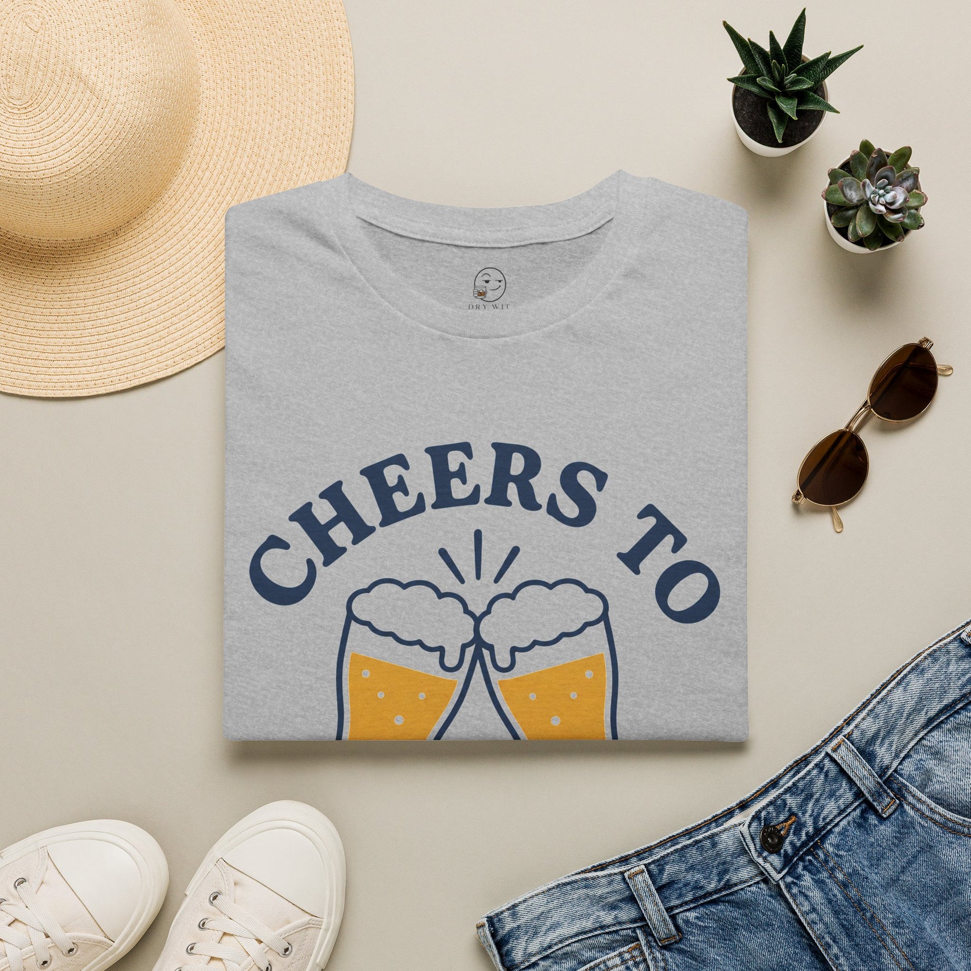 Cheers to Lowering Expectations Shirt, Sport Grey, folded flat lay styled with a straw hat, sunglasses, sneakers and denim