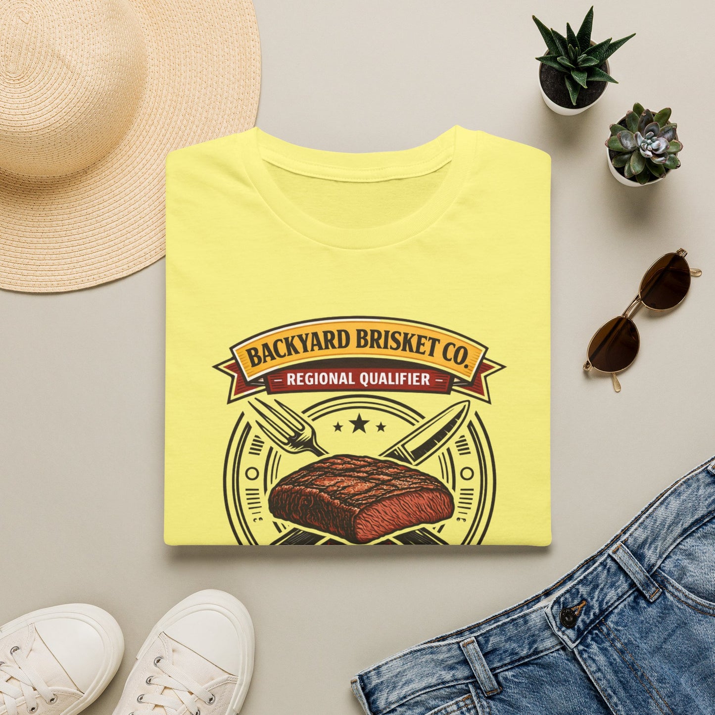 Backyard Brisket Co. Shirt, Cornsilk pale yellow, folded flat lay styled with a straw hat, sunglasses, sneakers and denim