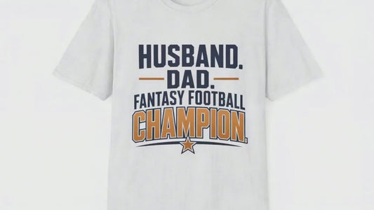Video of a man standing proudly holding up a small trophy with a big grin, wearing a white t-shirt with navy and amber text reading Husband Dad Fantasy Football Champion